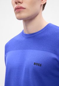 Kék kötött pulóver, amely textúrált vízszintes csíkkal, kerek nyakkivágással és fekete "BOSS" logóval rendelkezik a bal mellrészen.