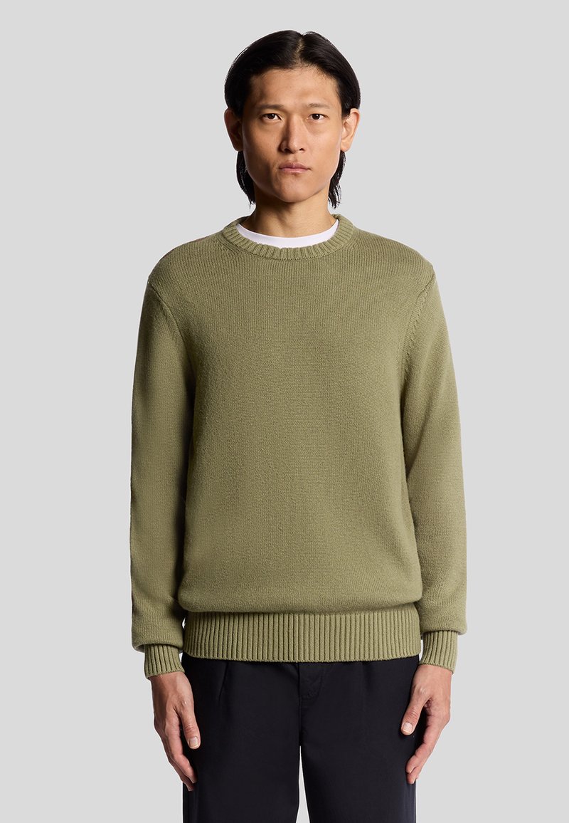 Pull en maille vert olive avec un col rond et des bords côtelés. Présente un tissu texturé et une coupe décontractée, porté par-dessus une chemise blanche.