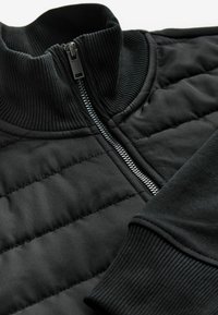Svart zip-up sweatshirt med hög ribbad krage, vadderade paneler och ribbade ärmar. Materialet ser slätt ut men är något texturerat.