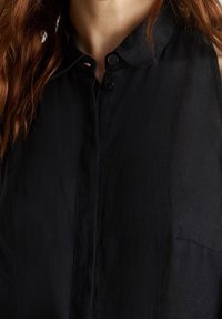 Femme avec de longs cheveux roux portant une chemise noire à boutons avec un col, découvrant la partie supérieure de la poitrine et le cou.