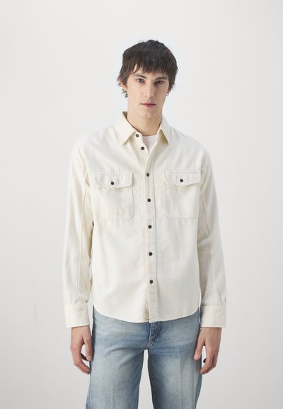 Camisa de manga larga color crema hecha de algodón, con dos bolsillos en el pecho, botones negros y un corte relajado, combinada con jeans de color azul claro.