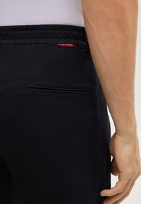 Czarne spodnie o fakturowanym wykończeniu, z elastycznym pasem i tylną kieszenią z patką. Widoczna czerwona metka z logo.