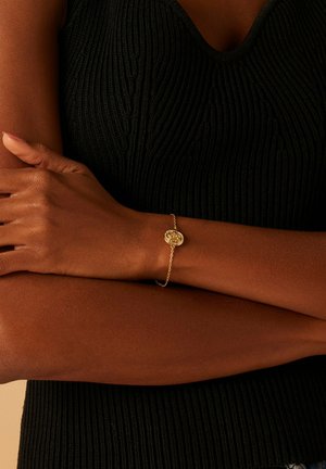 Bracelet en or avec un motif de rose circulaire, relié par une chaîne délicate. Porté sur un haut sombre et texturé. La teinte de la peau est chaude.