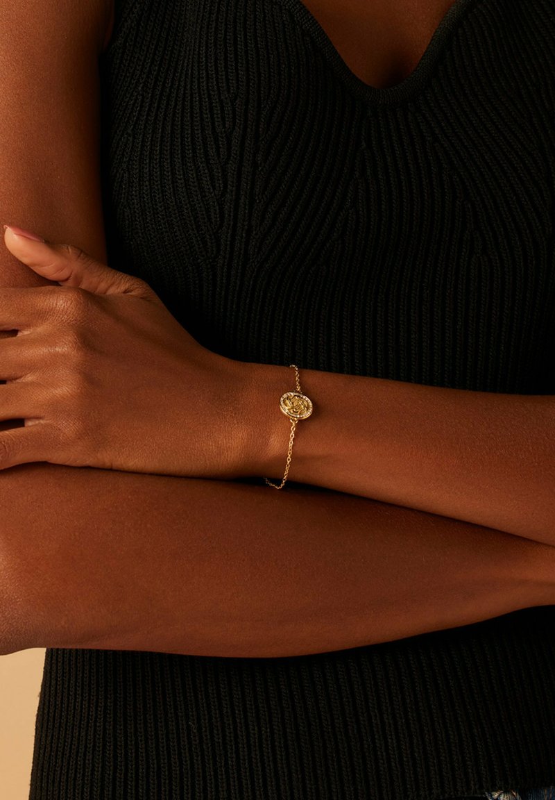 Gouden armband met een circulair rozenontwerp, verbonden door een delicate ketting. Draagbaar op een donkere, gestructureerde top. Huidskleur is warm.