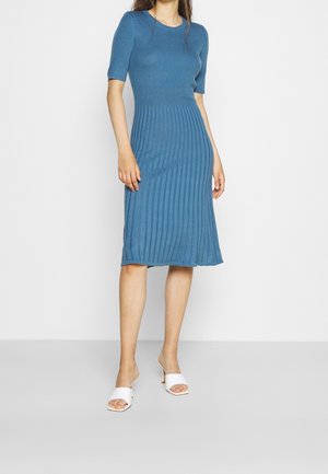 Kobieta ubrana w niebieską prążkowaną dzianą sukienkę o długości do kolan z krótkimi rękawami oraz białe sandały na obcasie z odkrytymi palcami, stojąca na jednolitym tle.