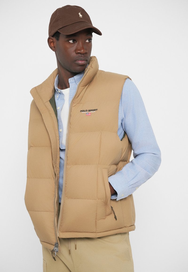 Gilet imbottito beige con colletto alto, chiusura con zip e tasche laterali. Indossato sopra una camicia azzurro chiaro con bottoni, accompagnato da un cappellino marrone.