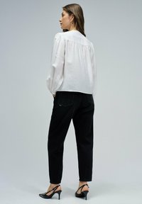 Blouse blanche à manches longues avec un dos plissé, associée à un jean noir taille haute et des sandales à talons noires. Texture de tissu lisse.