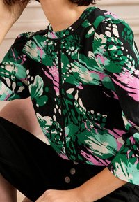 Blusa multicolore con sfondo nero e motivi astratti in verde, rosa e bianco; scollatura rotonda e maniche a sbuffo.