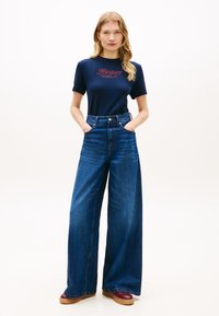 Γυναίκα στέκεται με τα χέρια στις τσέπες, φορώντας μπλε μπλουζάκι Tommy Hilfiger, φαρδιά μπλε τζιν παντελόνια και μπορντό παπούτσια σε απλό φόντο.