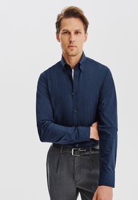 Granatowa koszula z długim rękawem o fakturowanej tkaninie, z kołnierzykiem zapinanym na guziki i dopasowanym kroju, zestawiona z szarymi spodniami i czarnym paskiem.