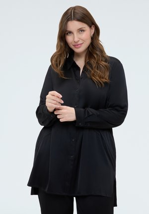 Junge Frau mit welligem braunem Haar, trägt ein lockeres schwarzes Hemd mit Knöpfen und schwarze Hose, steht vor einem schlichten hellen Hintergrund.