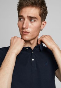 Jūrinės tamsiai mėlynos spalvos polo marškiniai su apykakle, papuošti dviem baltais mygtukais ir mažo išsiuvinėjimo detale. Modelis laiko apykaklę abiem rankomis.