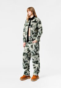 Conjunto de chaqueta y pantalones impresos en tonos de verde y gris. Presenta un corte relajado, cierre de cremallera y calzado marrón con suela gruesa.