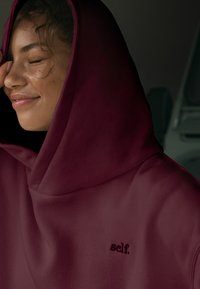 Burgundy huppari, joka on valmistettu pehmeästä materiaalista ja jossa on yksinkertainen brodeerattu logo "self." edessä. Pehmeä rakenne, rento malli.