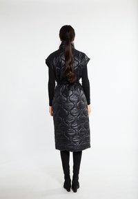 Gilet nero trapuntato con colletto alto e maniche corte, fissato con una cintura in vita, abbinato a leggings neri aderenti e tacchi.