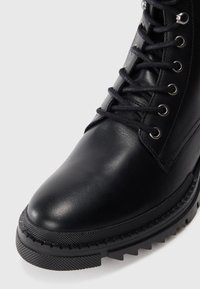 Botín de cuero negro con puntera redondeada, suela texturada y ojales contrastantes con cordones oscuros. Detalles de costura a lo largo de los lados.