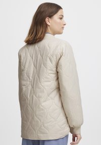 Beige quiltad jacka med en avslappnad passform, med rund hals och elastiska manschetter, som visar en mjuk textur och vågformat mönster.