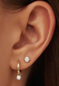 Boucle d'oreille en or avec un cristal transparent suspendu en bas et un petit cristal en stud au-dessus, sur un gros plan d'une oreille.