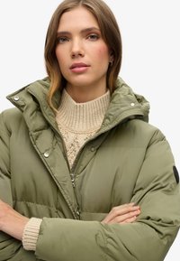 Chaqueta acolchada verde aceituna con un cuello alto y cierre de cremallera, llevada sobre un suéter de punto color crema, que presenta un logotipo negro en la manga.