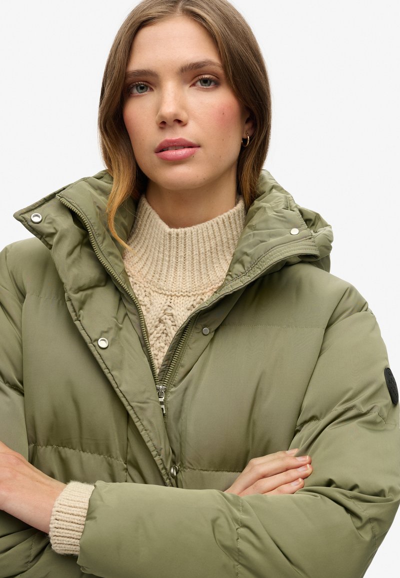 Olivgrüne Pufferjacke mit hohem Kragen und Reißverschluss, getragen über einem cremefarbenen Strickpullover, der ein schwarzes Logo am Ärmel aufweist.