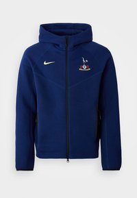Marineblå zip-up hoodie lavet af blød stof, med en sort lynlås, Nike-logo i gul og et broderet emblem på brystet.