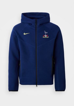 Marineblå zip-up hoodie lavet af blød stof, med en sort lynlås, Nike-logo i gul og et broderet emblem på brystet.