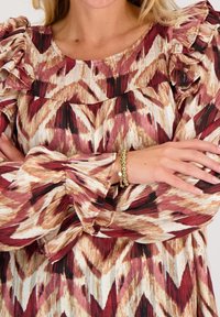 Patroonblouse met ruffelmouwen, versierd met geometrische vormen in tinten van rood, bruin en wit. Inclusief een gouden armband om de pols.