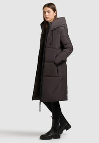 Lang, mørkebrun pufferjakke med hætte, der har en lynlås, sidelommer og en glat, quiltet tekstur. Bæres med sorte støvler og leggings.