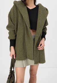 Olivgrön, fluffig, oversized kappa med huva, med stora knappar och svarta ärmdekorationer, i kombination med en svart crop top och ljus kjol.