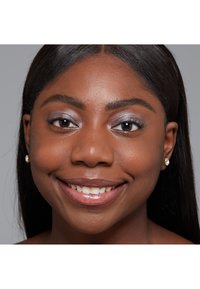 Close-up portrait of a person with dark skin, straight black hair, silver eyeshadow, and small pearl earrings, smiling against a gray background.