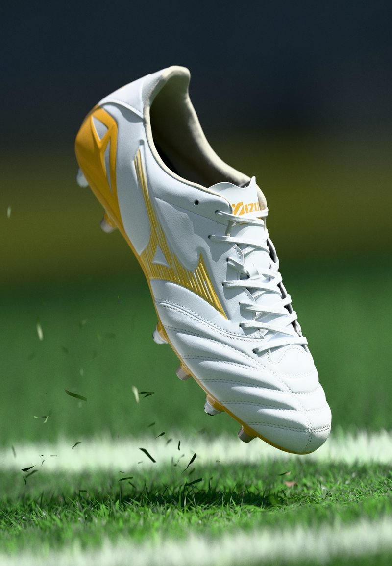 Άσπρο και κίτρινο παπούτσι ποδοσφαίρου να σηκώνει γρασίδι σε πράσινο γήπεδο με λευκές γραμμές ορίων στο φόντο.