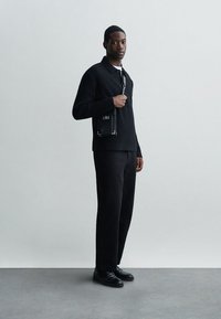 Μαύρος πλεχτός πουλόβερ με γιακά, συνδυασμένος με μαύρο παντελόνι ίσιας γραμμής και μαύρα παπούτσια. Μια μικρή μαύρη τσάντα χιαστί ολοκληρώνει το όλο look.