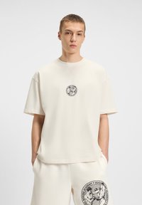 Lys beige oversized t-shirt med korte ærmer, der har et sort cirkulært grafisk print på brystet og en afslappet kant.