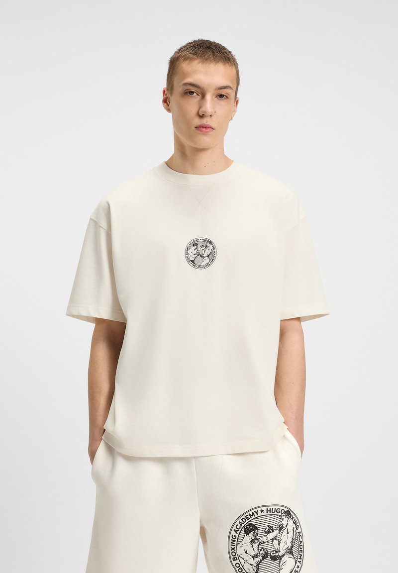 Lys beige oversized t-shirt med korte ærmer, der har et sort cirkulært grafisk print på brystet og en afslappet kant.