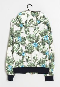 Hooded jack met een witte achtergrond, voorzien van groene bladeren en blauwe bloemen. Inclusief een marineblauwe geribbelde onderrand en manchetten met een gouden ritsdetail.