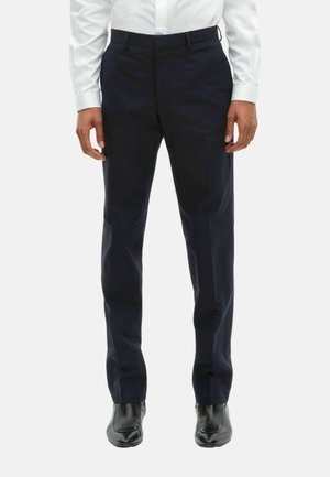 Homme portant un pantalon noir formel, des chaussures de ville noires et une chemise blanche à manches longues, se tenant devant un fond blanc.