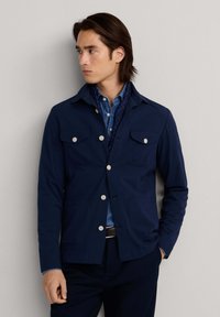 Homme portant une veste bleue marine boutonnée sur une chemise en denim avec une écharpe à pois, debout devant un fond gris clair, regardant vers la gauche.
