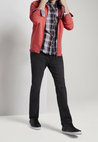 Hoodie rouge à fermeture éclair porté sur une chemise à carreaux boutonnée, associé à un jean noir et des baskets noires avec des accents gris. Design simple et décontracté.