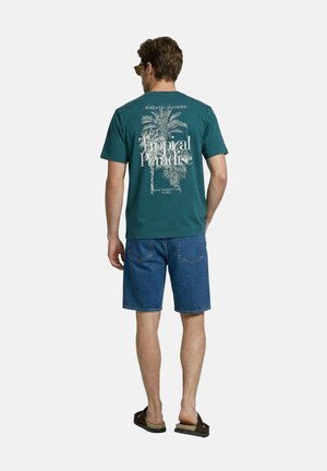 Man wearing teal Tropical Paradise T-shirt, blue denim shorts, brown sandals, and sunglasses facing away on plain background.