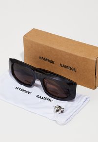 Occhiali da sole neri rettangolari con lenti marroni, accompagnati da una pochette in microfibra bianca con il logo "SAMSNØE" e una scatola di cartone marrone.