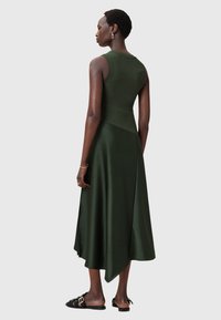 Abito senza maniche di colore verde scuro con corpetto a costine, gonna in satin fluida e orlo asimmetrico. Il modello indossa sandali a scivolo e gioielli minimi.