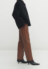 Pull noir oversize avec un col montant et des détails de boutons, associé à un pantalon en cuir marron à jambes droites et des escarpins à talons fins noirs.