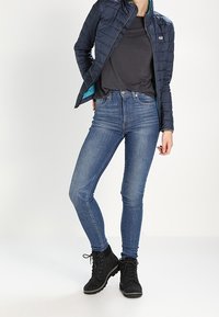 Marinblå kviltad jacka med tealfärgad foder, svart t-shirt, blå slim fit-jeans och svarta vandringsskor. Leggingspassform och funktionella designelement.