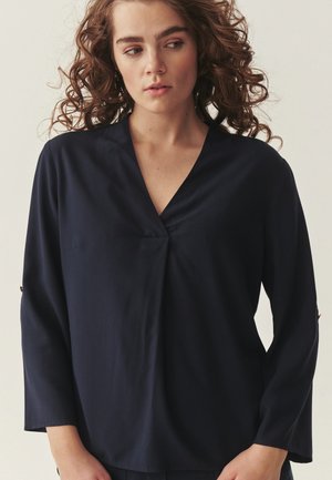 Femeie cu păr creț purtând o bluză bleumarin cu guler în V și mâneci lungi, privind ușor spre stânga, pe un fundal simplu.