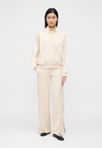 Bež pulover z naročjem z zadrgo do polovice, stranskimi žepi in temnejšimi bež poudarki na rokavih. Hlače širokih nog z bočnimi razporki v enaki barvi.