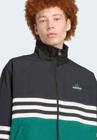 Must musta ja roheline Adidas jope, millel on lukuga sulgemine, kõrge krae ja kolm valget triipu rinna kohal roheliste aktsentidega.