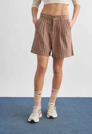 Άτομο που φοράει σορτς με ψηλή μέση και καρό σχέδιο, με τα χέρια στις τσέπες, μπεζ κάλτσες Nike και λευκά αθλητικά παπούτσια, στέκεται σε μπλε χαλί.