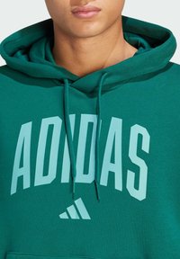 Sweatshirt vert Adidas avec capuche, orné d'un grand texte et logo blancs. Fabriqué en tissu doux avec des cordons de serrage et une poche avant.