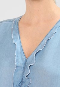 Helles blaues Denim-Bluse mit V-Ausschnitt, ausgestattet mit Rüschenkanten und dezenten Nähdetails. Weiche Textur und lässiges Design.