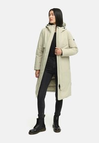 Lichtbeige gewatteerde jas met capuchon, met een rits aan de voorkant, een trekkoord onderaan en zijzakken, gedragen over zwarte jeans en laarzen.